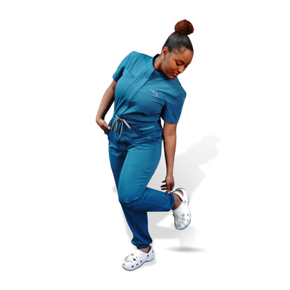 Woman modeling a teal scrub romper, standing on one leg to show the fit and style of the garment.